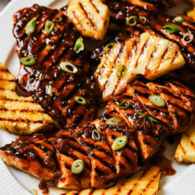 A white plate with Grilled Pineapple Chicken breasts topped with a dark glaze and sliced green onions, served alongside smoky grilled pineapple wedges.