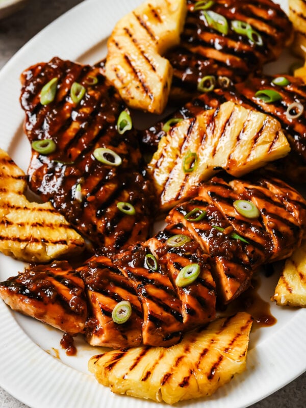 A white plate with Grilled Pineapple Chicken breasts topped with a dark glaze and sliced green onions, served alongside smoky grilled pineapple wedges.