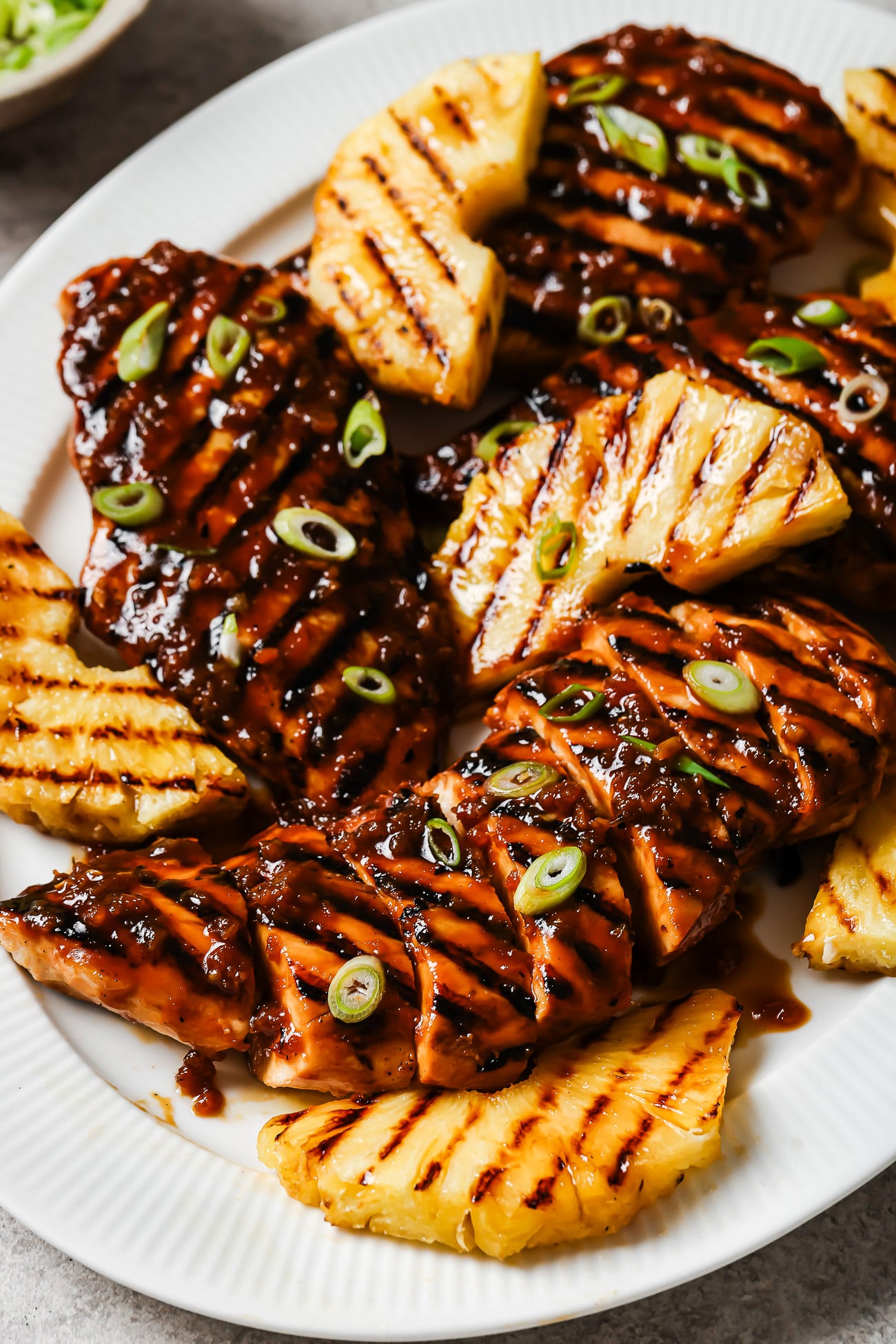 A white plate with Grilled Pineapple Chicken breasts topped with a dark glaze and sliced green onions, served alongside smoky grilled pineapple wedges.