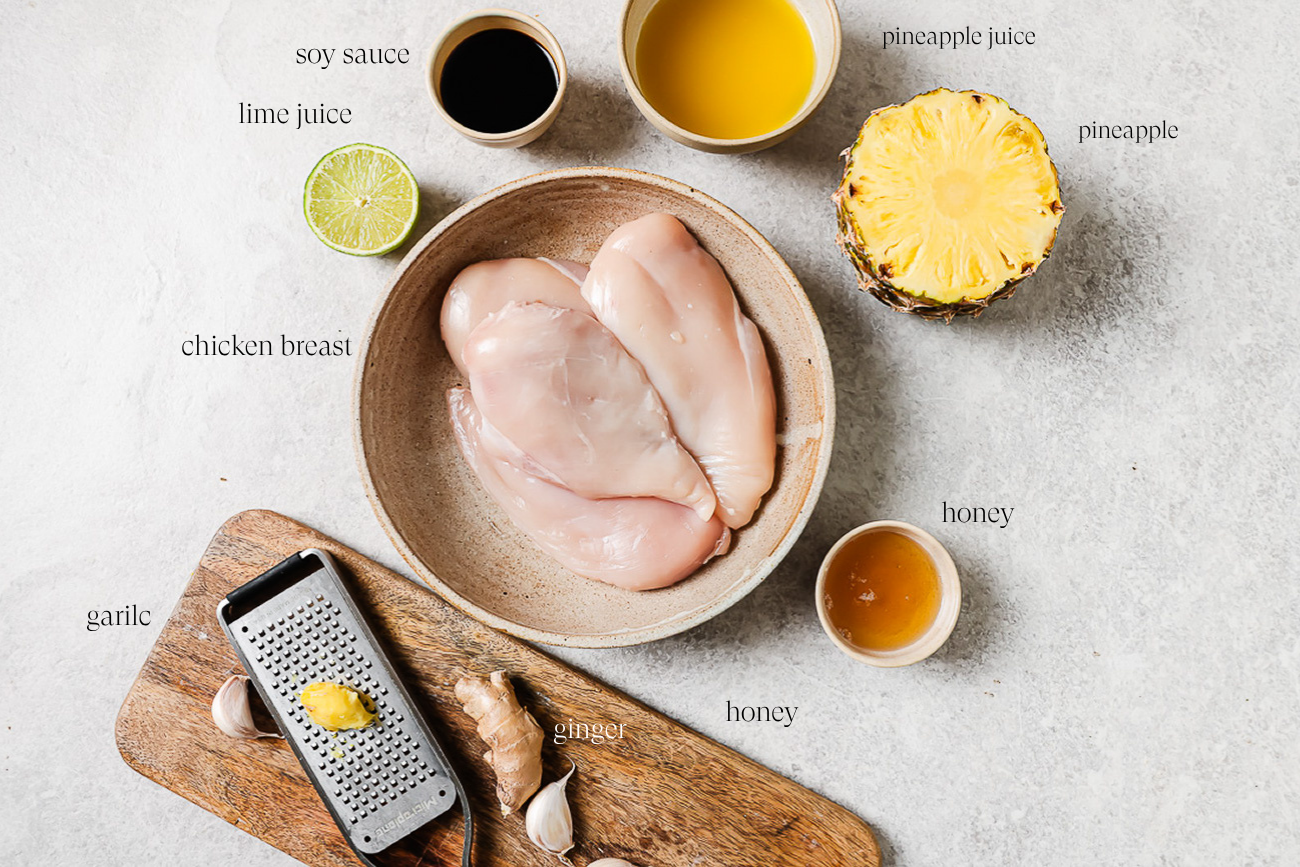 A bowl with raw chicken breasts, surrounded by labeled ingredients for Grilled Pineapple Chicken: soy sauce, lime juice, pineapple juice, half a pineapple, honey, garlic, and grated ginger on a cutting board.