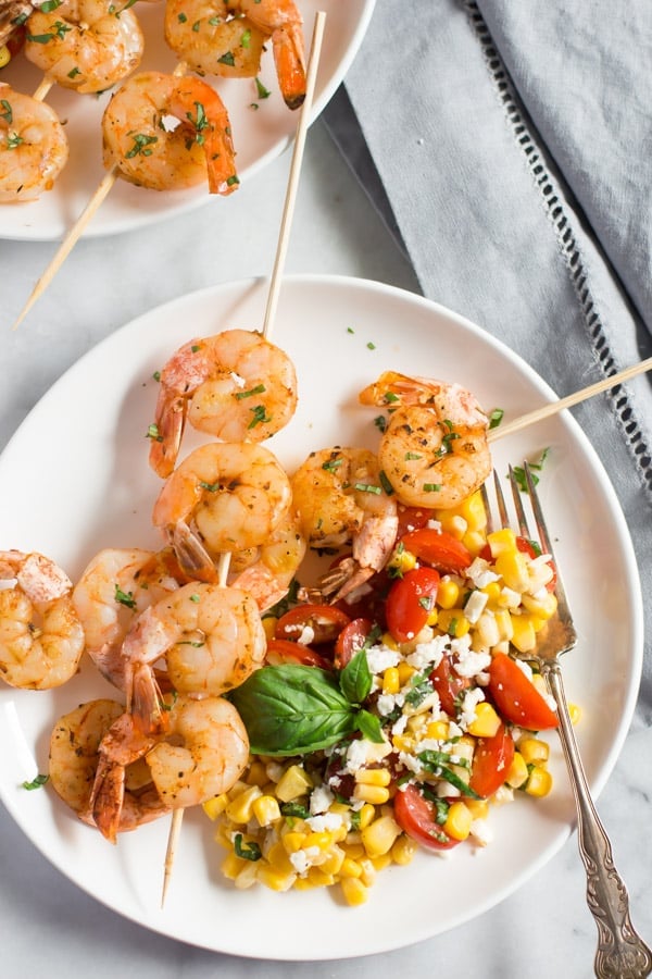 A white plate with two shrimp skewers on top of a corn salad.