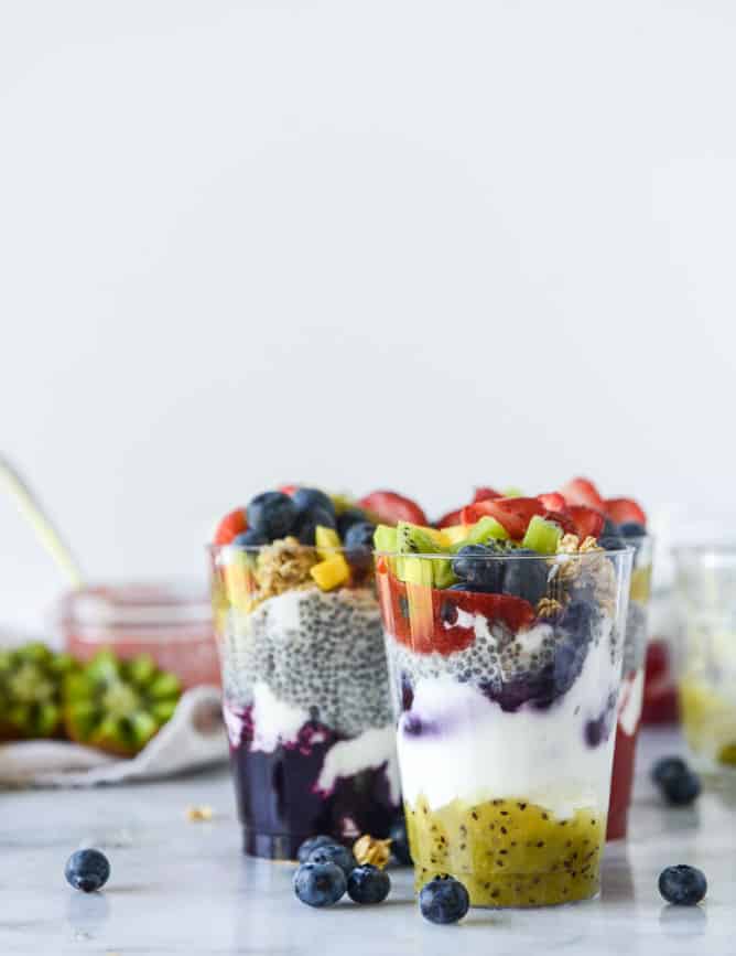Two Rainbow Syrup Bottomed Chia Pudding Parfaits in clear cups topped with kiwi, berries, and granola.
