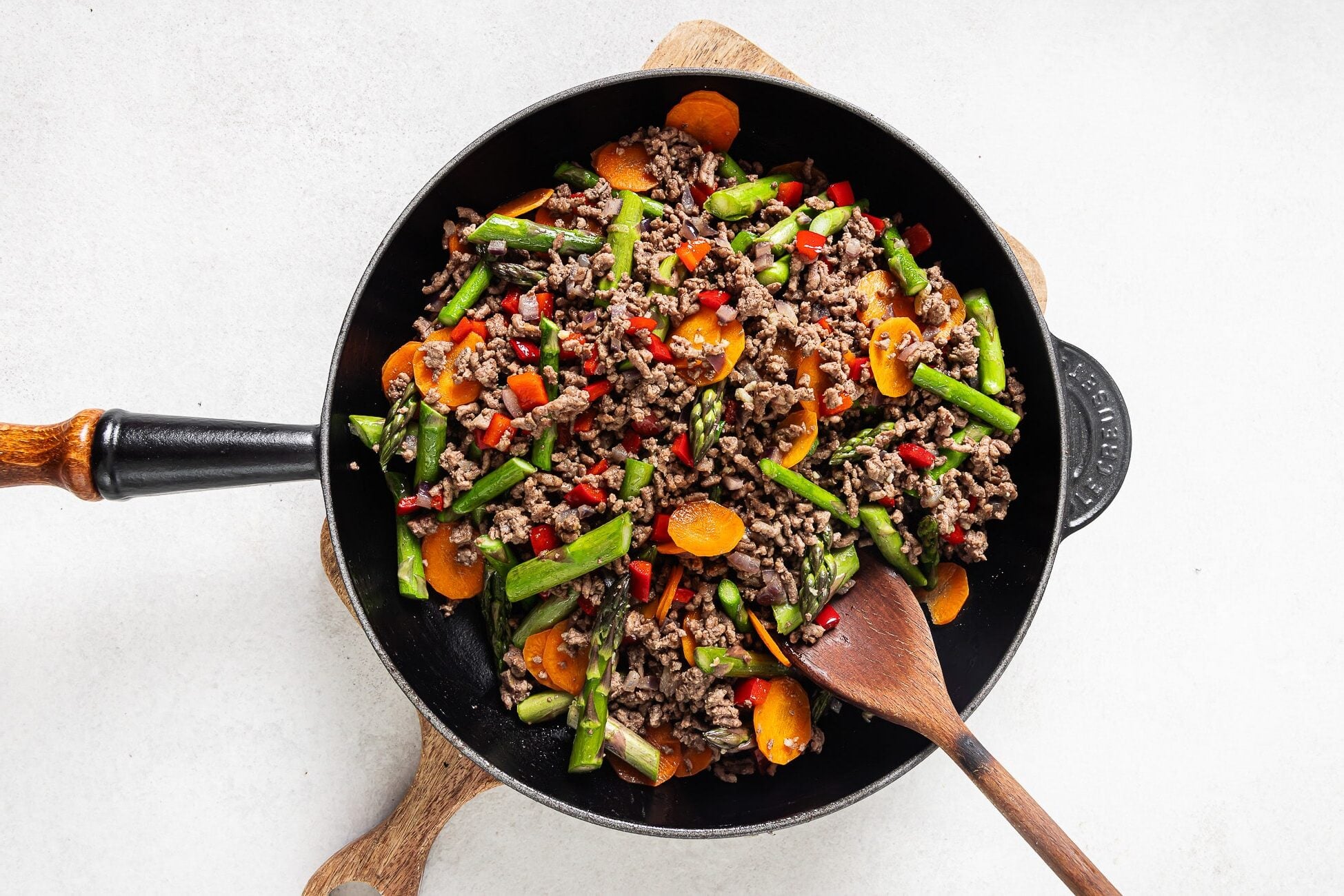 A black skillet filled with Ground Beef with Vegetables—sliced carrots, chopped green beans, red bell peppers, and asparagus—sits on a wooden board with a wooden spoon resting inside.