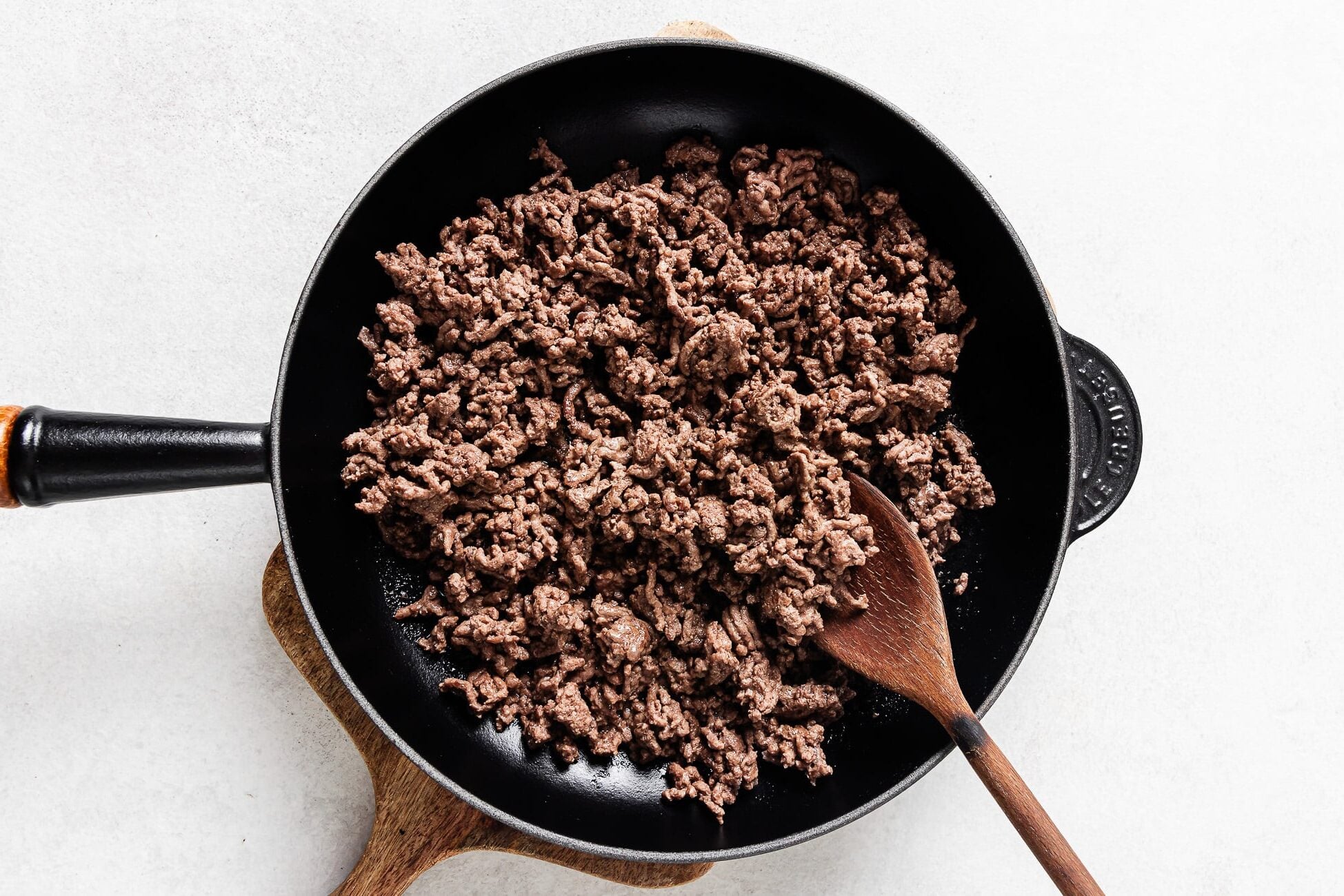 Cooked ground beef with vegetables in a black skillet, stirred with a wooden spoon, is set atop a wooden cutting board against a white background.