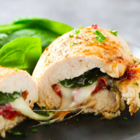A Spinach Stuffed Chicken Breast, filled with spinach, melted cheese, and sun-dried tomatoes, is sliced and served on a black plate with fresh spinach leaves in the background.