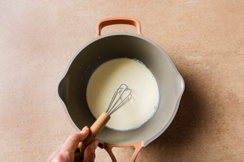 A hand whisks a creamy white mixture in a large gray saucepan with tan handles, prepping the batter for a Low-carb Almond Flour Chocolate Cake on a light brown countertop.