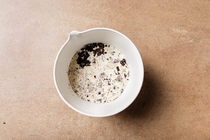A white bowl with a spout containing heavy cream and chopped dark chocolate pieces, perfect for making a Low-carb Almond Flour Chocolate Cake, sits on a light brown countertop.