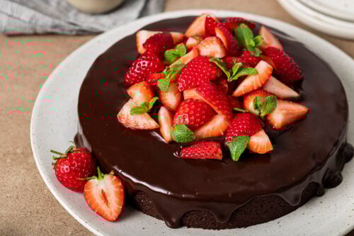 A round Almond Flour Chocolate Cake topped with glossy chocolate ganache, fresh sliced strawberries, and mint leaves, served on a white plate. A whole and a halved strawberry rest on the plate beside the cake.