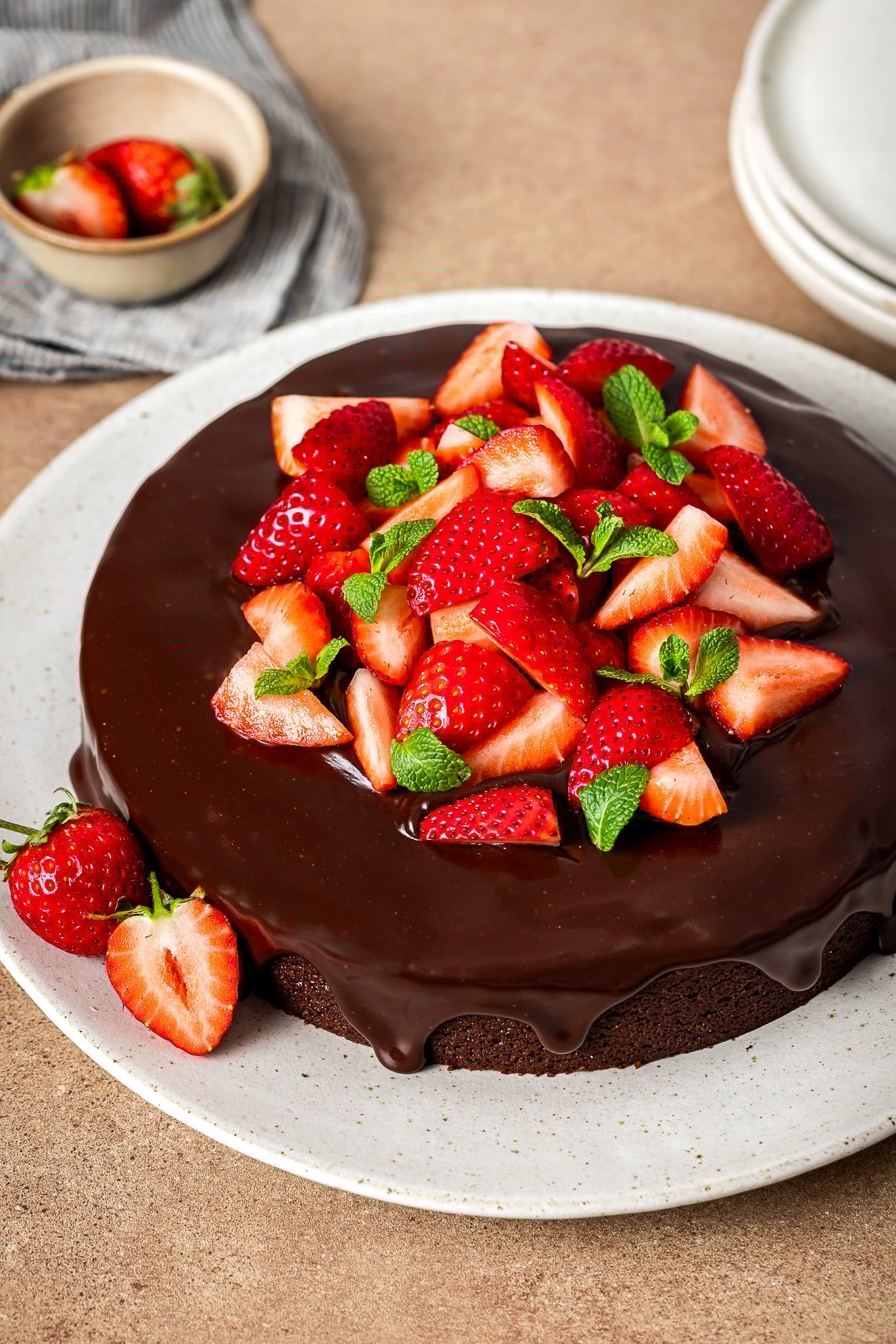 A Almond Flour Chocolate Cake topped with glossy chocolate ganache, fresh sliced strawberries, and mint leaves, served on a white plate with extra strawberries in a small bowl in the background.