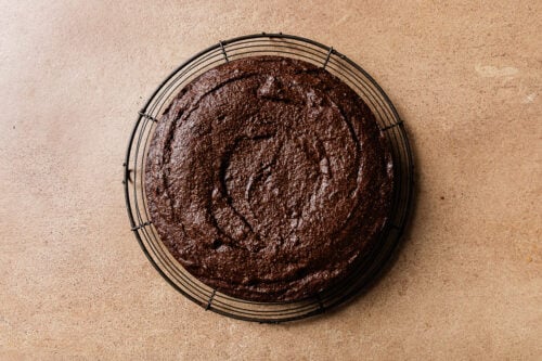A round, unfrosted chocolate cake cools on a black wire rack, placed on a light brown textured surface.