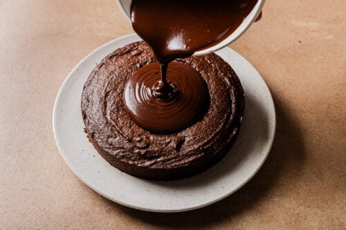 A bowl of glossy chocolate ganache is being poured onto the center of a round Almond Flour Chocolate Cake on a white plate, set on a brown surface.