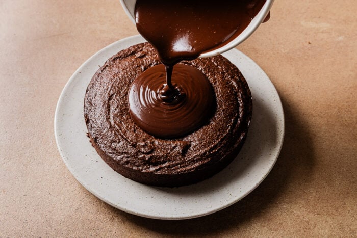 A bowl of glossy chocolate ganache is being poured onto the center of a round Low-carb Almond Flour Chocolate Cake on a white plate, set on a brown surface.