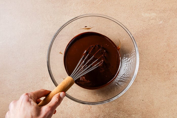 A hand using a wooden-handled whisk to mix melted chocolate for a low-carb almond flour chocolate cake in a clear glass bowl on a beige countertop.