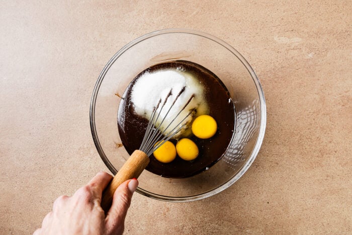 A hand holds a whisk in a glass bowl containing Low-carb Almond Flour Chocolate Cake batter, three raw eggs, and a mound of sugar, all on a light brown countertop.