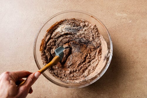 A hand uses a spatula to mix dry ingredients, including cocoa powder, sugar, and almond flour for this chocolate Cake, in a clear glass bowl on a brown countertop.