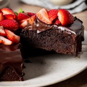 A rich, low-carb almond flour chocolate cake topped with glossy chocolate ganache and fresh sliced strawberries sits on a plate. A slice has been cut and partially removed, revealing the cake’s moist interior.