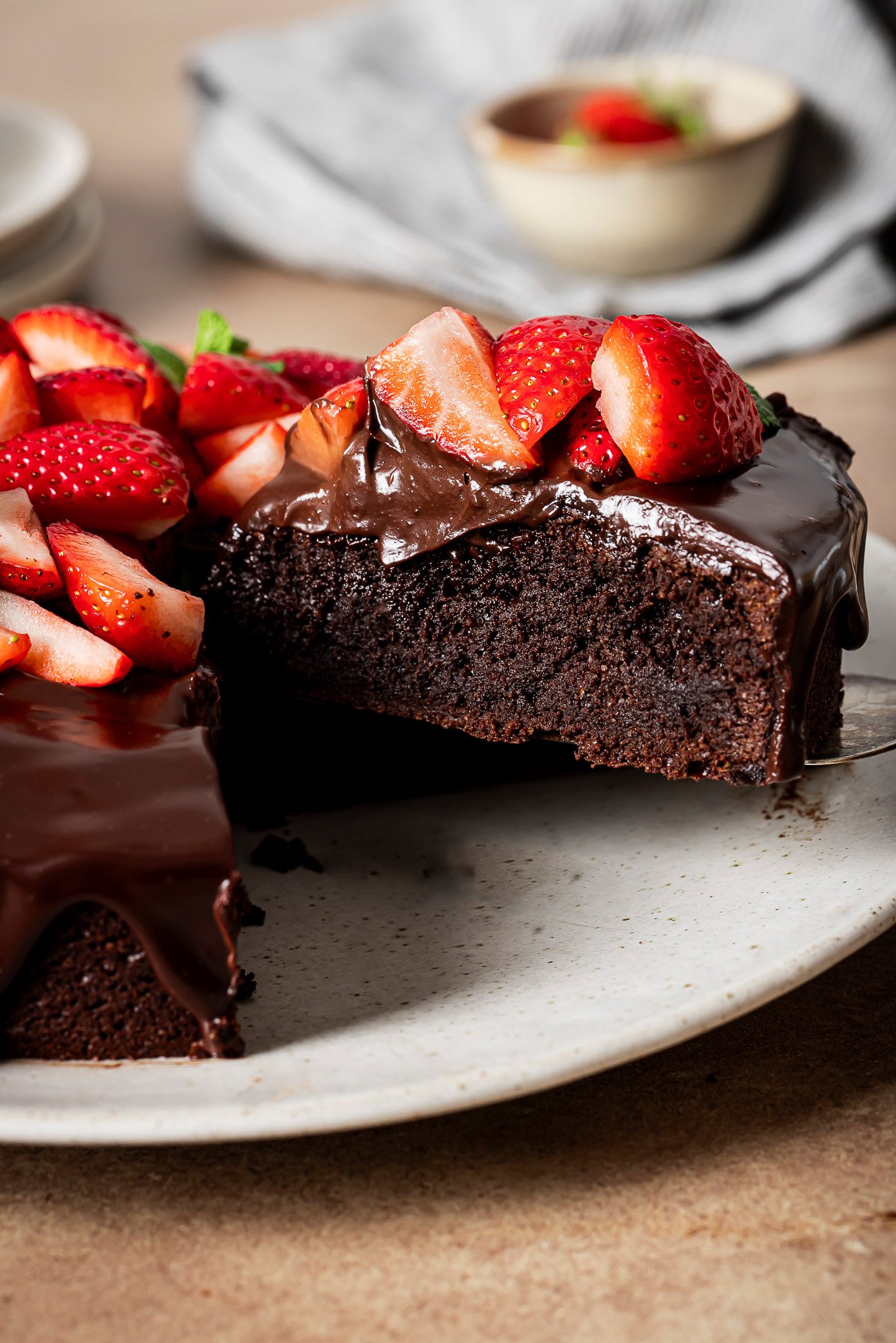 A rich, low-carb almond flour chocolate cake topped with glossy chocolate ganache and fresh sliced strawberries sits on a plate. A slice has been cut and partially removed, revealing the cake’s moist interior.