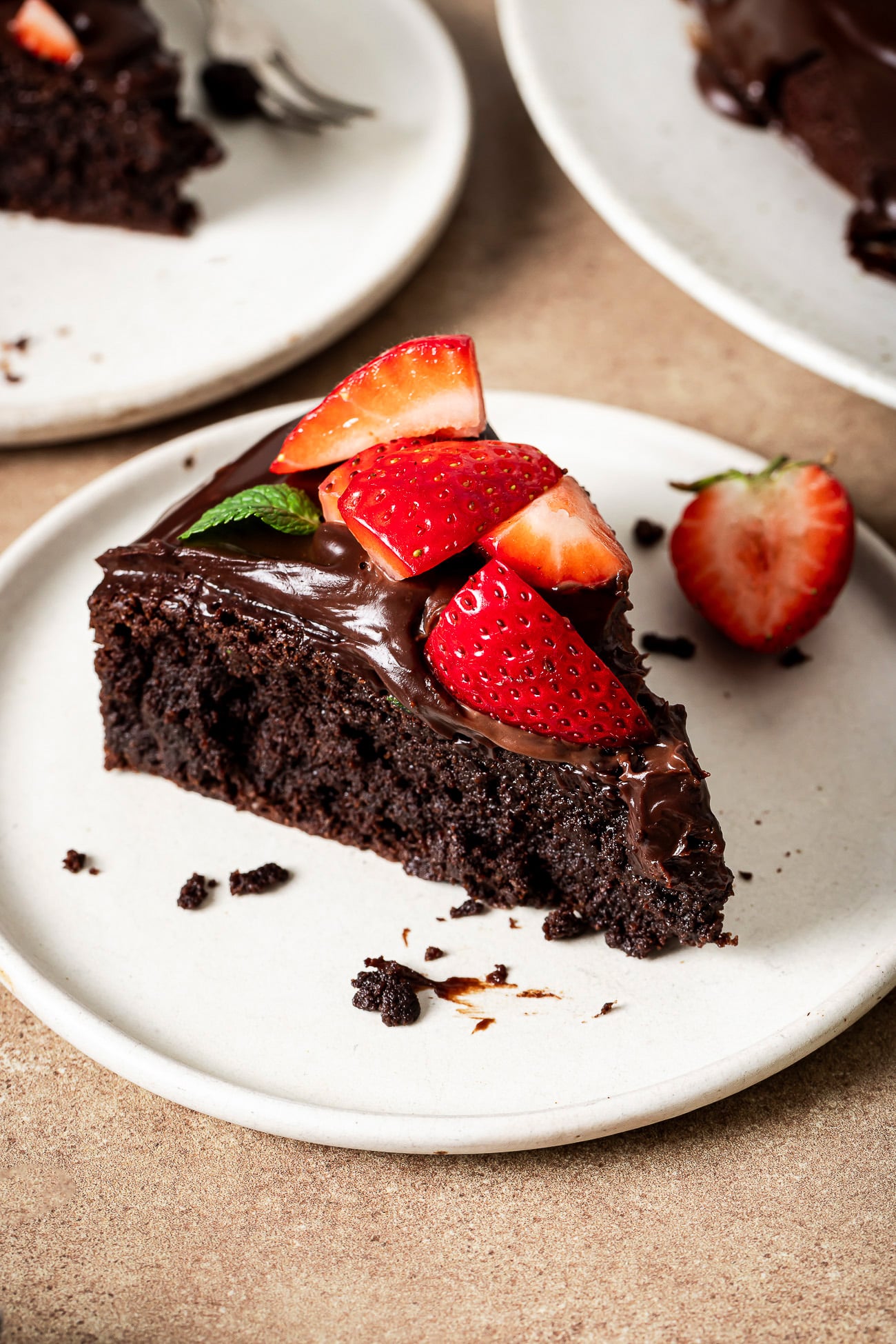 A slice of low-carb Almond Flour Chocolate Cake with glossy chocolate frosting, topped with fresh strawberry pieces and a mint leaf, on a white plate. Another plate with cake is visible in the background.