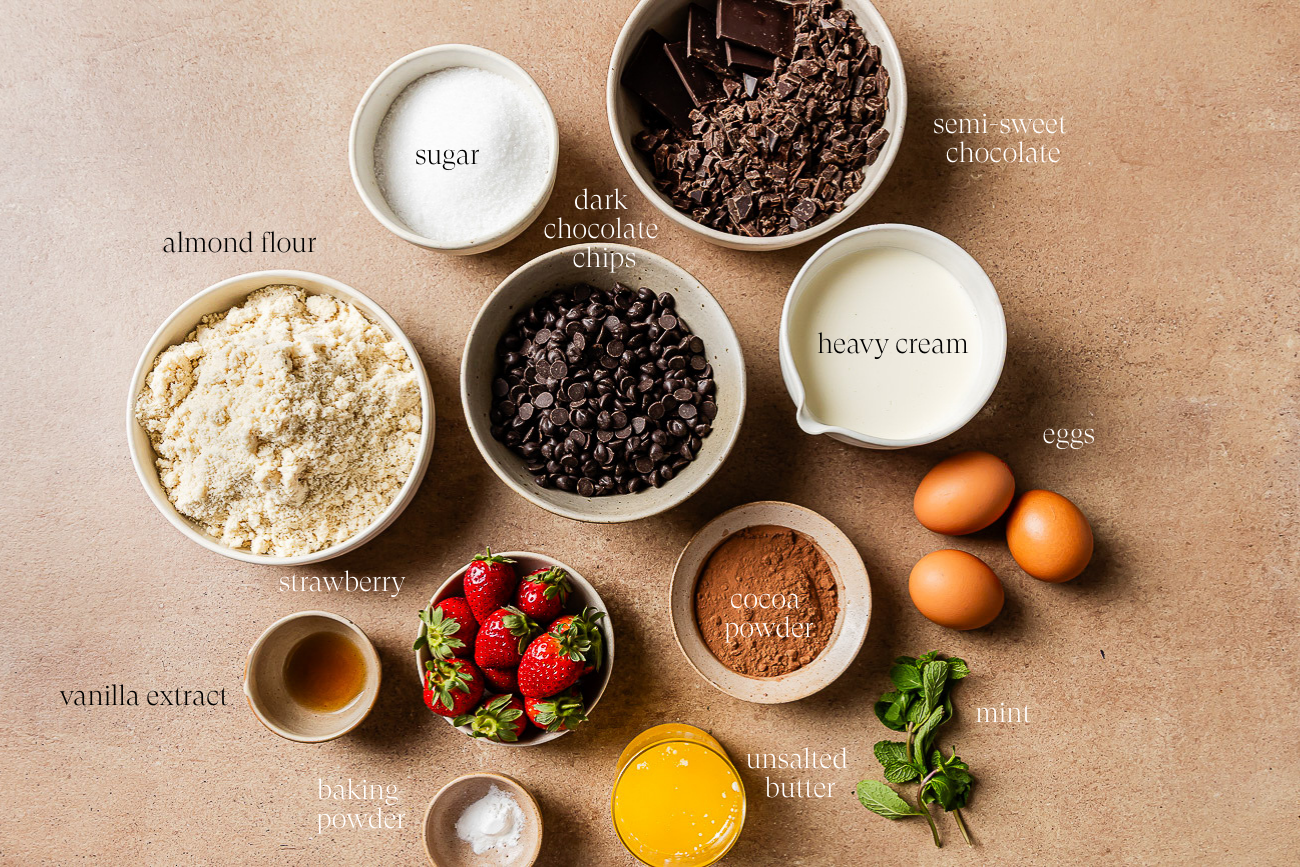 A flat lay of baking ingredients for a Low-carb Almond Flour Chocolate Cake, featuring almond flour, sugar, dark chocolate chips, semi-sweet chocolate, heavy cream, eggs, strawberries, vanilla extract, cocoa powder, unsalted butter, baking powder, and mint.