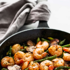 A black skillet filled with cooked shrimp, asparagus, and sliced garlic sits on a white surface with a gray cloth nearby. The Shrimp Asparagus Recipe is written above, highlighting the delicious combination of fresh seafood and crisp vegetables.