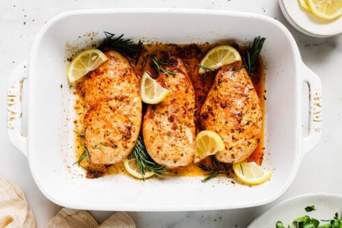 Three seasoned and baked Lemon Rosemary Chicken breasts in a white baking dish, garnished with lemon wedges and sprigs of fresh rosemary. Some juices are visible in the bottom of the dish.
