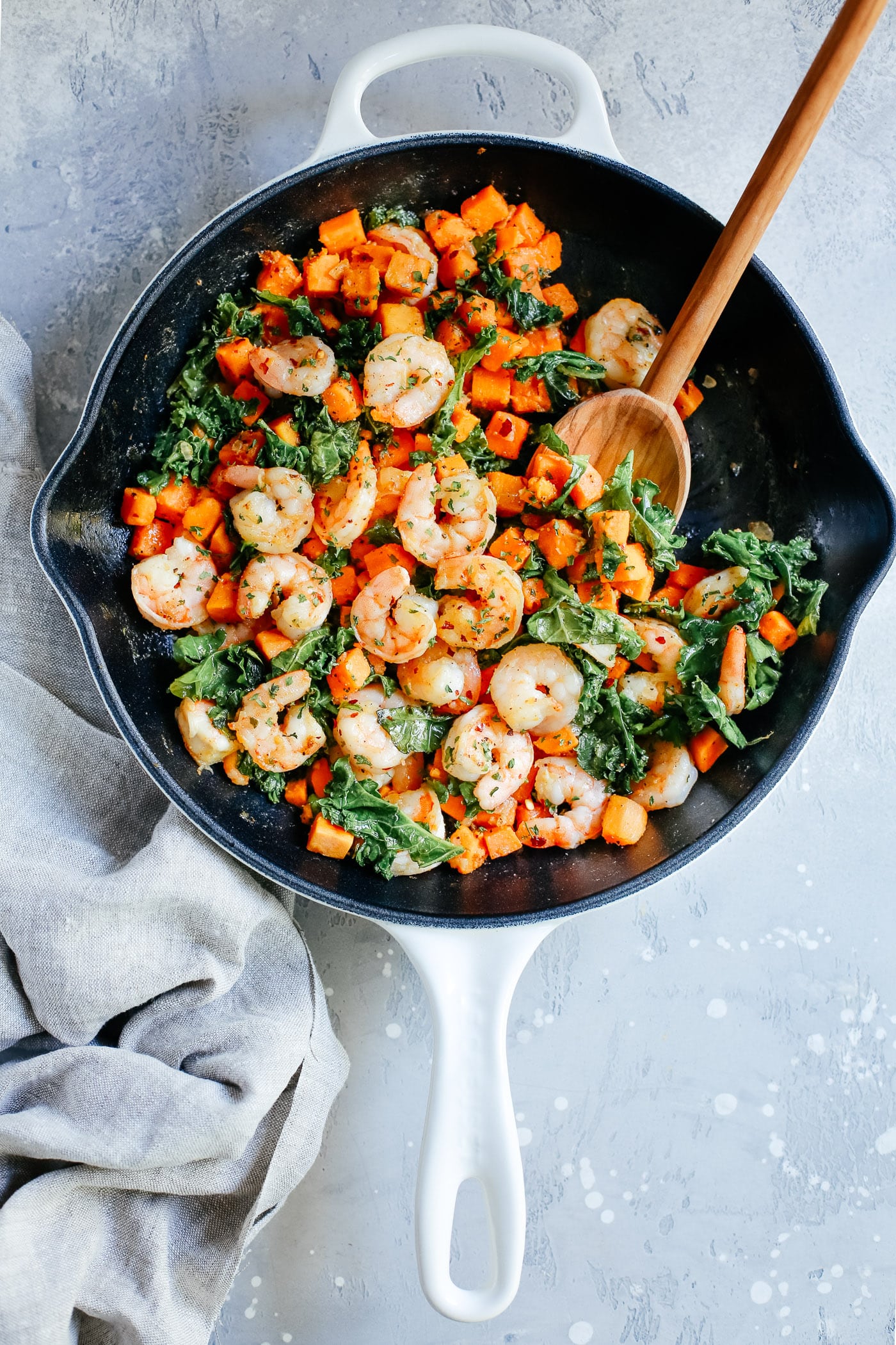 A white skillet with shrimp, kale, and sweet potatoes with a spoon inside.