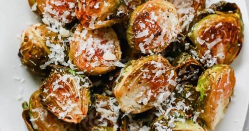 Close-up of Roasted Brussel Sprouts halves on a white plate, topped with grated Parmesan cheese. The Roasted Brussel Sprouts have crispy, browned edges and are finished with a light sprinkle of cheese.