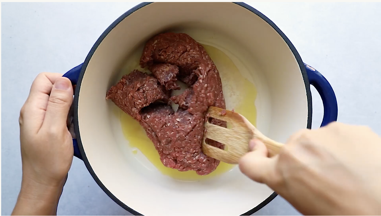 A person uses a wooden spatula to cook raw ground beef for a Ground Beef Taco Salad in a white pot with blue handles on a light surface.