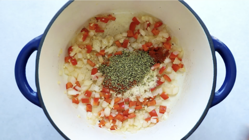 Chopped onions, red bell peppers, and seasonings—including dried herbs and paprika—are cooking in a white pot with blue handles, creating a flavorful base perfect for a hearty Ground Beef Taco Salad.