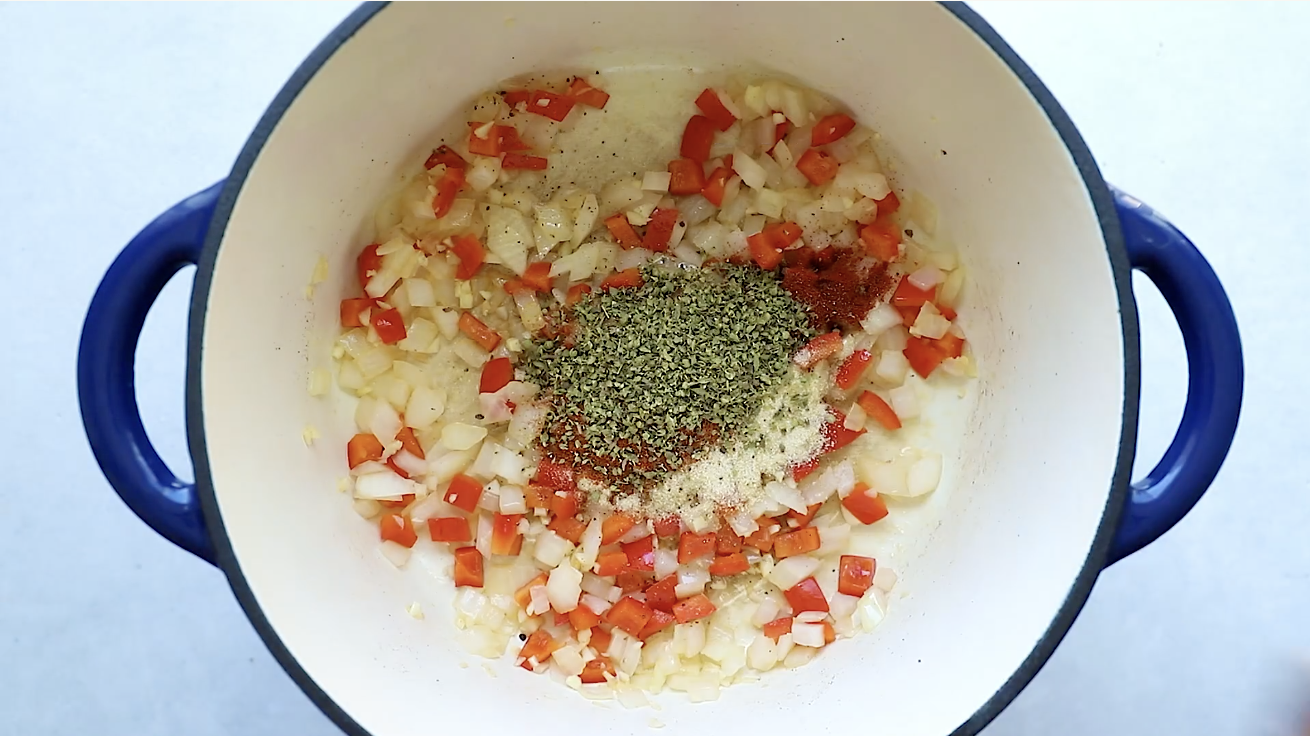 Chopped onions, red bell peppers, and seasonings—including dried herbs and paprika—are cooking in a white pot with blue handles, creating a flavorful base perfect for a hearty Ground Beef Taco Salad.
