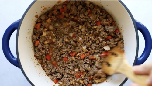 Ground beef, chopped onions, and red bell peppers are being cooked and stirred in a large white pot with a wooden spoon, forming the savory base for a delicious Ground Beef Taco Salad.