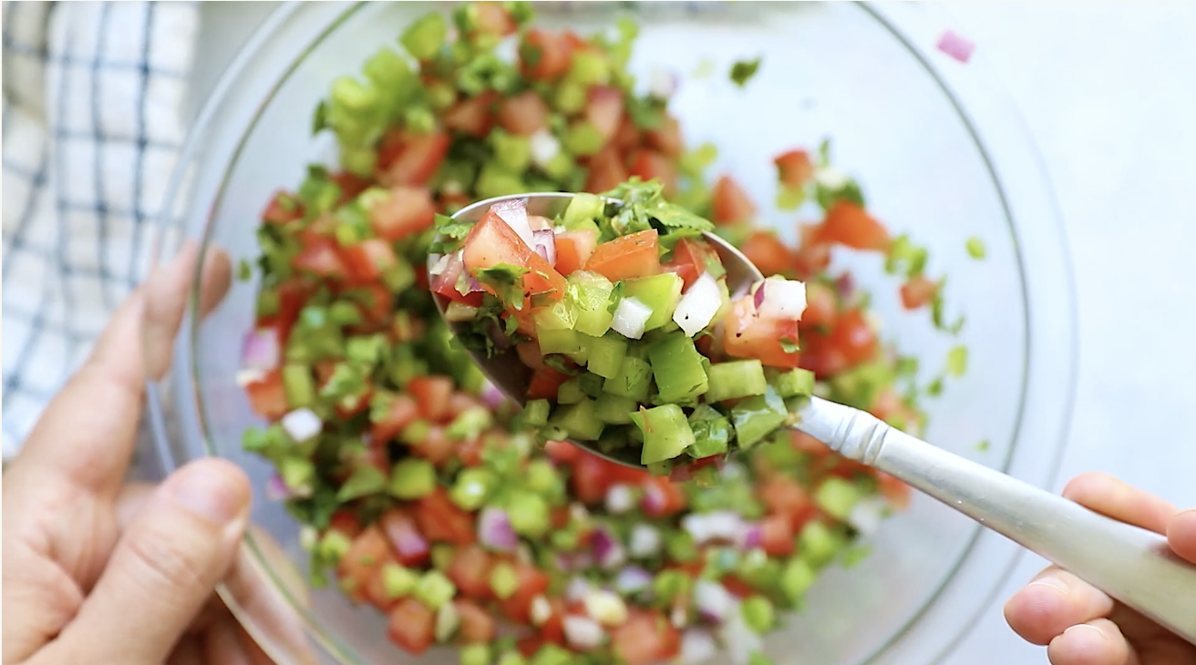 A close-up of a spoonful of fresh salsa with diced tomatoes, onions, green peppers, and cilantro over a glass bowl filled with the same colorful mixture, perfect as a topping for Ground Beef Taco Salad.