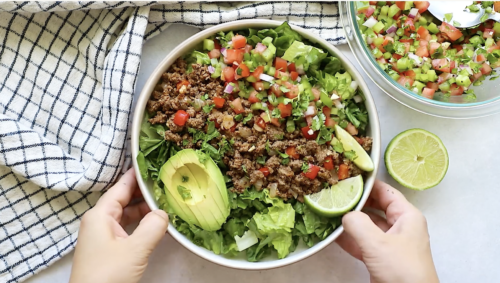 A bowl of Ground Beef Taco Salad with chopped lettuce, avocado slices, lime wedges, and salsa. Two hands hold the bowl beside a checkered cloth, with extra salsa and a halved lime on the side.