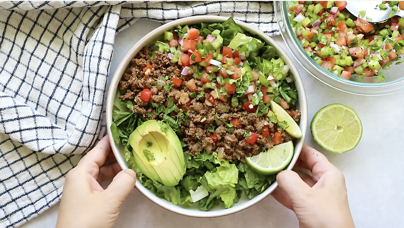 A bowl of Ground Beef Taco Salad with chopped lettuce, avocado slices, lime wedges, and salsa. Two hands hold the bowl beside a checkered cloth, with extra salsa and a halved lime on the side.