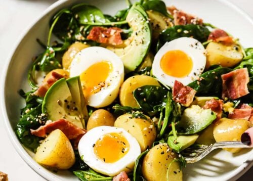 A bowl of salad featuring halved soft-boiled eggs, avocado slices, baby potatoes, spinach, crispy bacon, and black pepper sits next to a cup of coffee and a glass of orange juice on a white table.