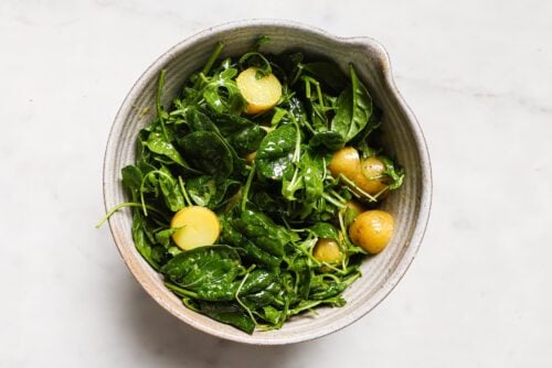 A ceramic bowl filled with fresh spinach leaves, arugula, and sliced small yellow potatoes on a white marble surface—an inviting Breakfast Salad Recipe idea.