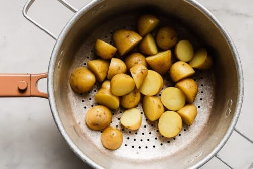 A metal colander with halved and quartered boiled baby potatoes, perfect for your next Breakfast Salad Recipe, sits on a white surface.