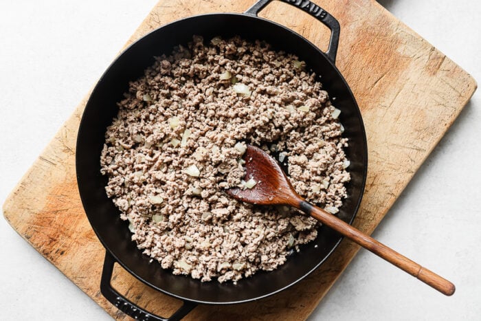 A black skillet filled with cooked ground turkey and chopped onions sits on a wooden cutting board, with a wooden spoon resting inside the pan—perfect for an easy ground turkey recipe.