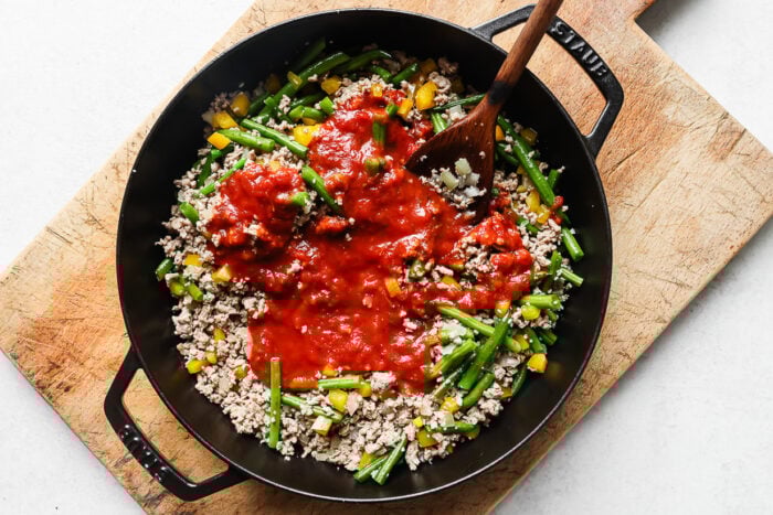 A black skillet on a wooden board contains ground urkey with cgreen beans, yellow bell peppers, onions, and tomato sauce being stirred with a wooden spoon.