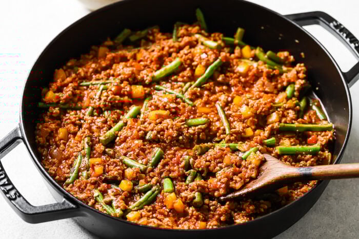 A black skillet filled with a tasty Ground Turkey recipe featuring ground meat, tomato sauce, chopped yellow bell peppers, and green beans, being stirred with a wooden spoon.