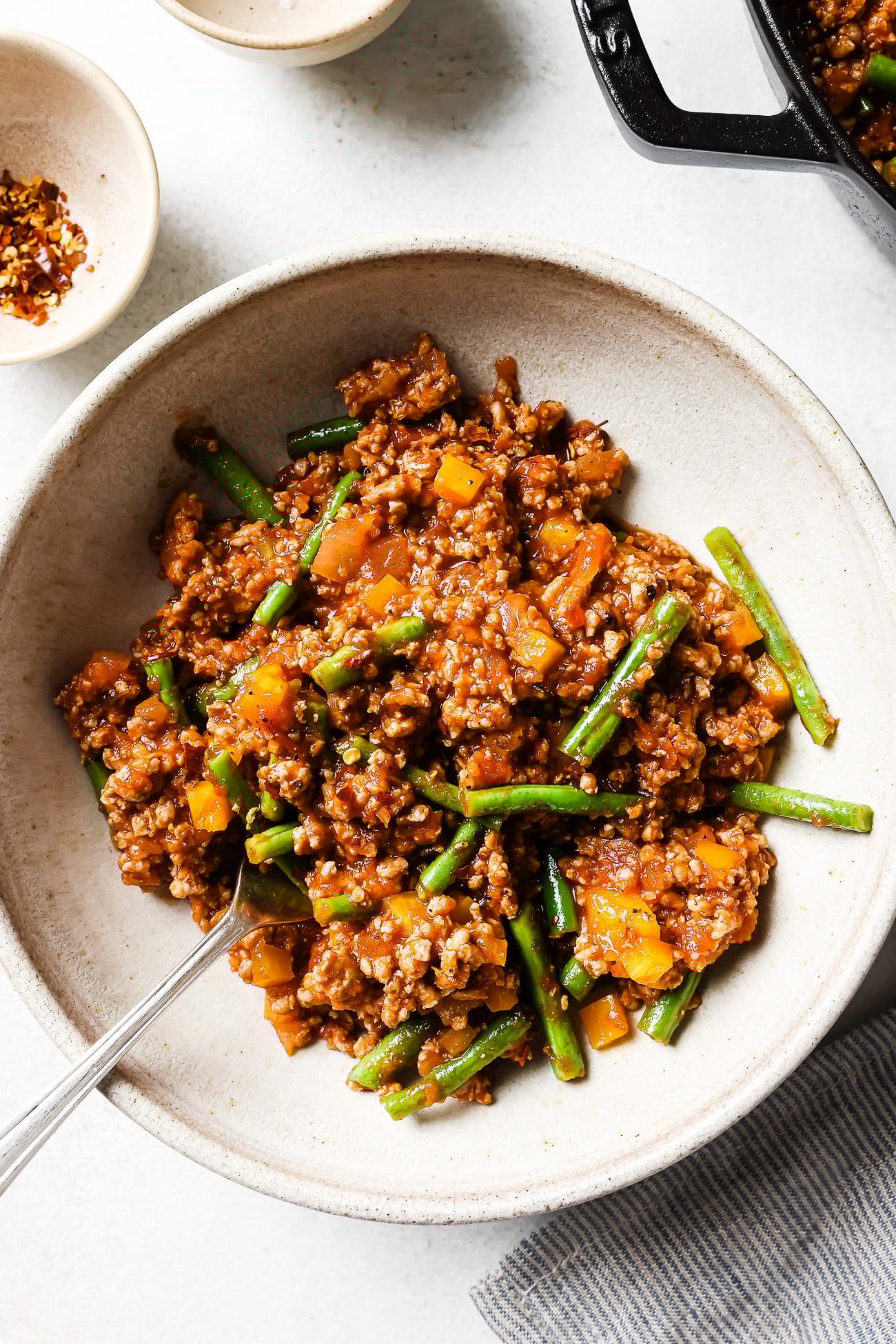 A bowl of ground turkey stir-fry with green beans and yellow bell peppers, served with a spoon. This colorful Ground Turkey recipe is saucy and vibrant, set on a light surface near spice bowls and a striped napkin.