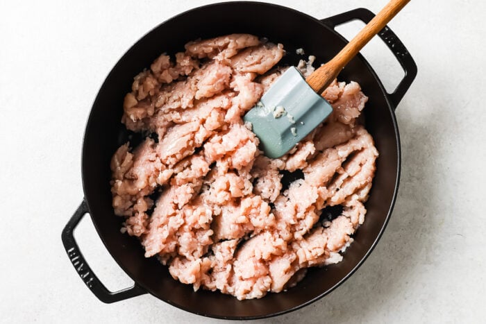 Raw ground chicken is being stirred with a gray spatula in a large black skillet on a light-colored surface, much like you would start this recipe.