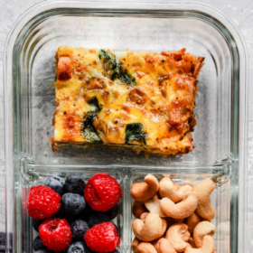 A glass meal prep container with three sections: a slice of baked egg casserole with spinach, mixed berries (raspberries and blueberries), and cashews. These Breakfast Meal Prep Bowls make mornings simple and nutritious.