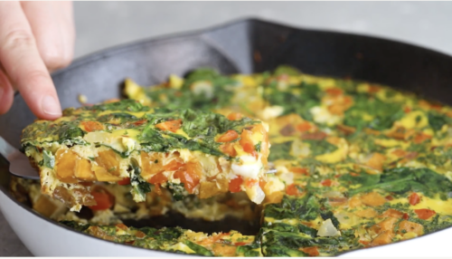 A close-up of a hand lifting a slice of vegetable frittata from a cast-iron skillet, showing colorful layers of spinach, bell peppers, and other vegetables—perfect for Breakfast Meal Prep Bowls.