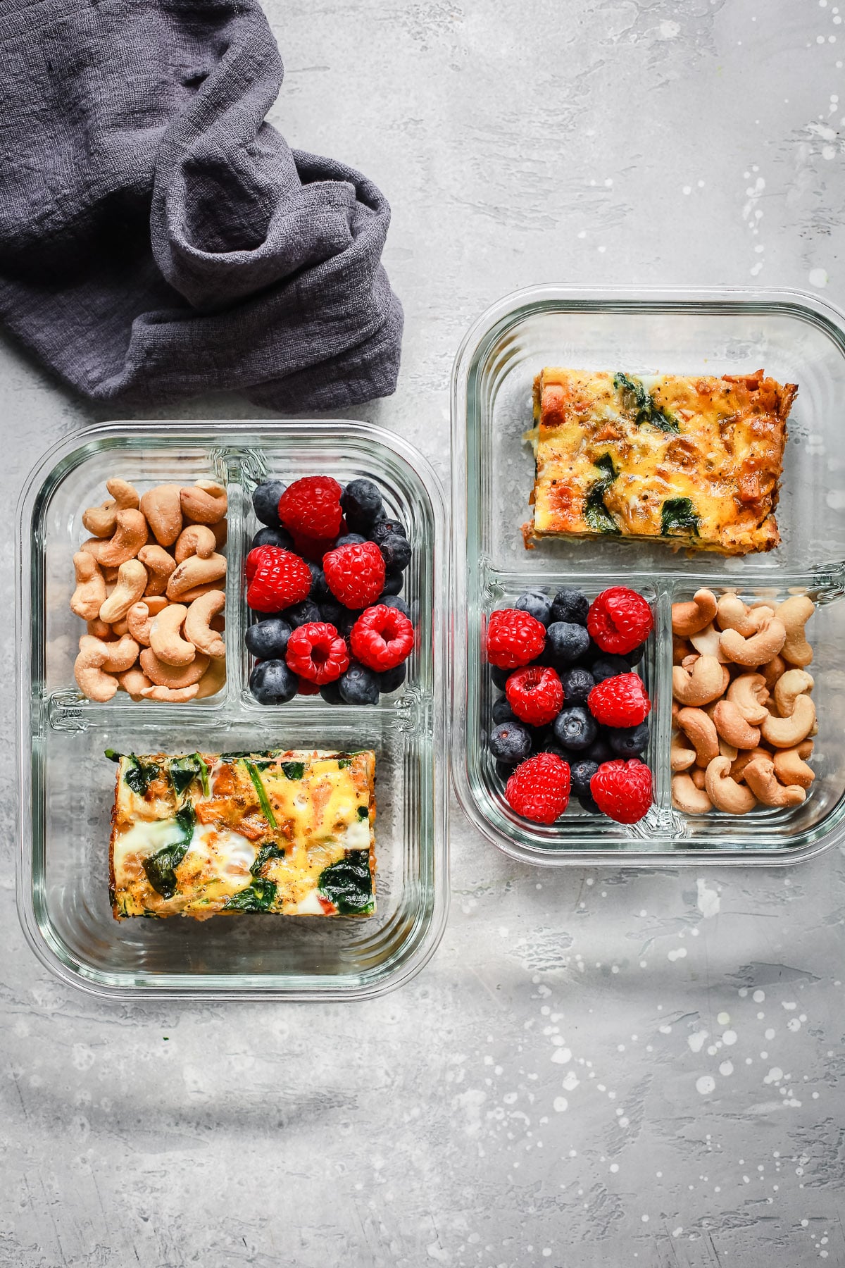 Two glass Breakfast Meal Prep Bowls with spinach egg casserole, cashews, raspberries, and blueberries are arranged on a light gray surface next to a folded dark gray cloth.