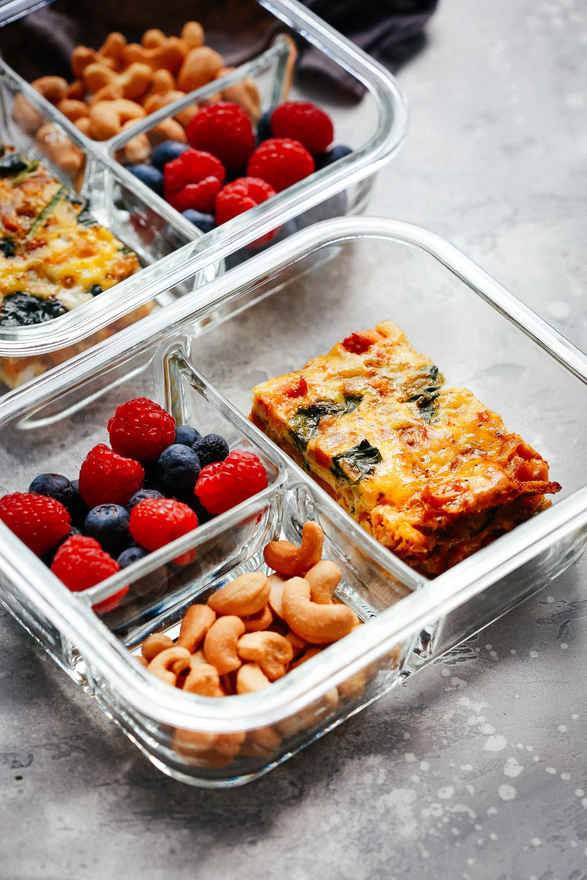 Two glass Breakfast Meal Prep Bowls, each divided into three sections with a piece of vegetable frittata, a handful of cashews, and a mix of fresh raspberries and blueberries, set on a light gray textured surface.