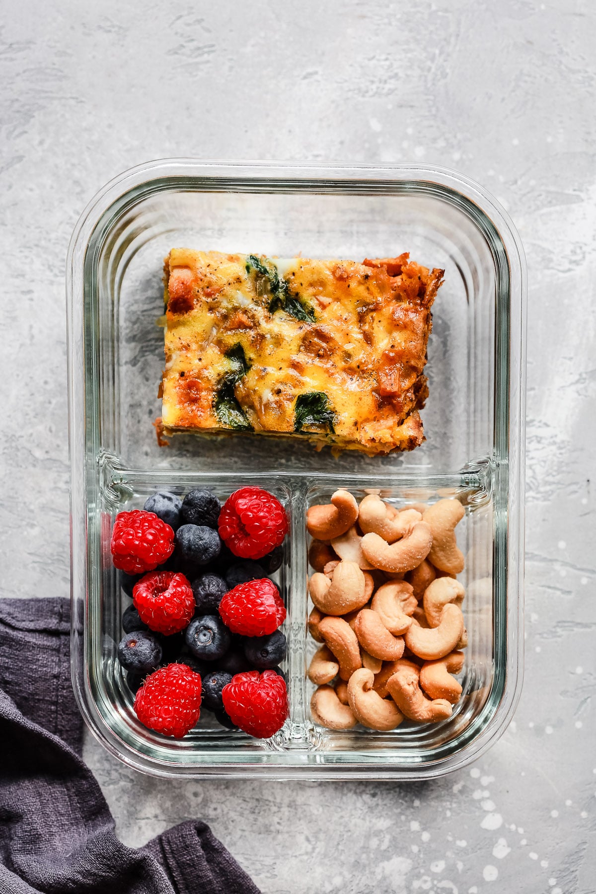 A glass meal prep container with three sections: egg casserole with spinach, fresh raspberries and blueberries, and a serving of cashews, on a gray textured surface&mdash;an ideal choice for Breakfast Meal Prep Bowls.