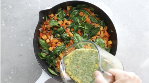 A hand pours a mixture of beaten eggs and herbs from a glass measuring cup into a skillet filled with sautéed spinach, orange bell peppers, and diced sweet potatoes—perfect for Breakfast Meal Prep Bowls.