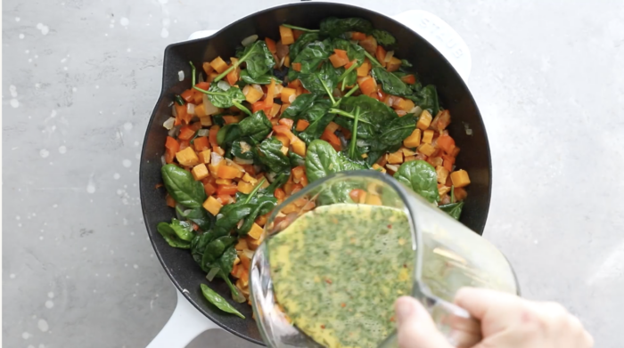 A hand pours a mixture of beaten eggs and herbs from a glass measuring cup into a skillet filled with sautéed spinach, orange bell peppers, and diced sweet potatoes—perfect for Breakfast Meal Prep Bowls.