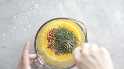 A hand whisks eggs with dried herbs and red pepper flakes in a glass measuring cup on a light gray countertop, perfect for Breakfast Meal Prep Bowls.